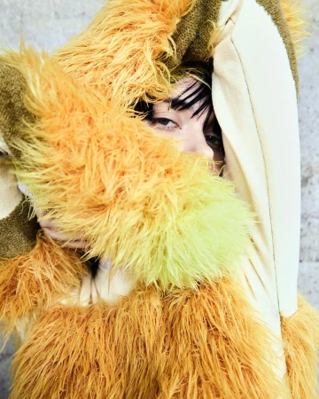 Close-up portrait of a person with brown hair partially obscured by a fuzzy, yellow, orange, and beige hooded jacket against a gray wall.