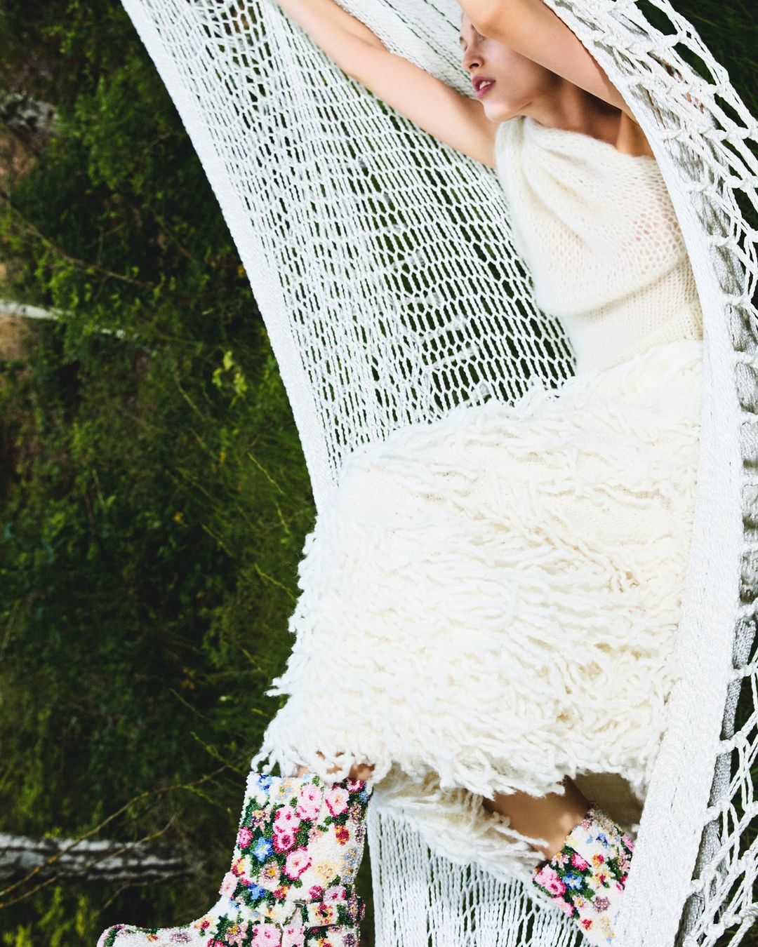 An adult female model relaxes in a white hammock outdoors, wearing a white knit dress and flower-print boots, surrounded by a lush green background.