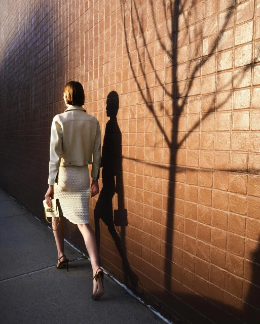 A woman in a skirt and jacket walks on a sidewalk next to a brick wall, with a tree and person shadow on the wall.