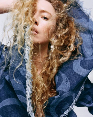 Close-up portrait of a woman with long curly blonde hair wearing a patterned blue denim hooded jacket.
