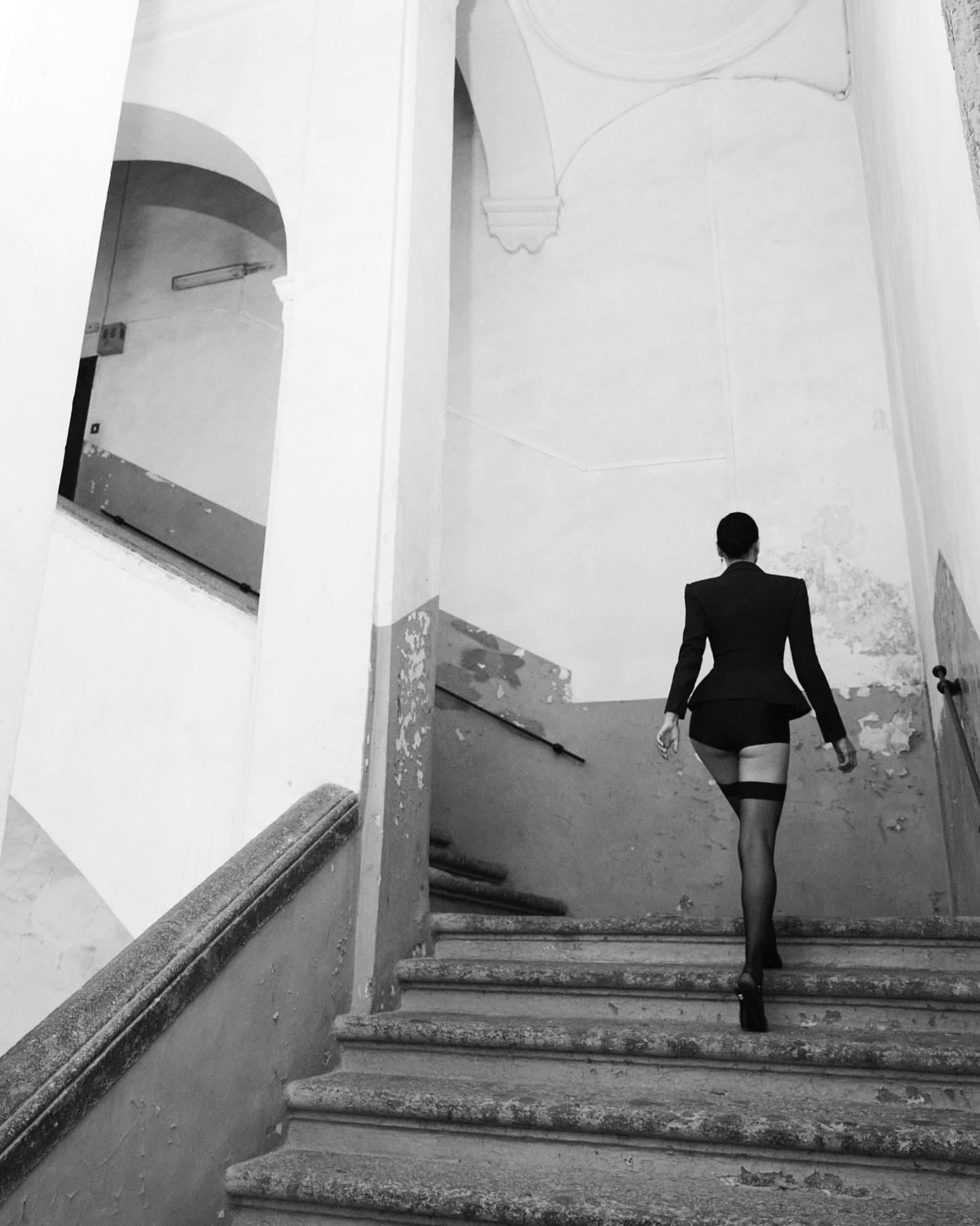 A woman in a suit jacket and stockings is seen from the back, ascending an old staircase in an indoor setting, captured in black and white.
