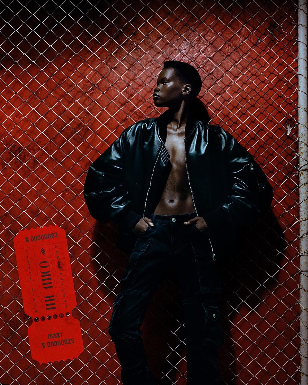 A stylish Black woman poses against a red wall and chain-link fence, wearing a black bomber jacket and dark jeans, shot in studio lighting.