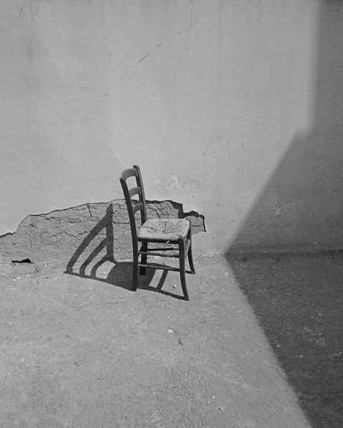 Monochrome photograph of a rustic wooden chair in a room corner, casting shadows on the floor and textured wall, creating contrast.