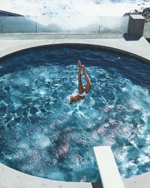 Overhead view of adult female diving into a circular pool with turquoise water and a diving board against a coastal landscape.