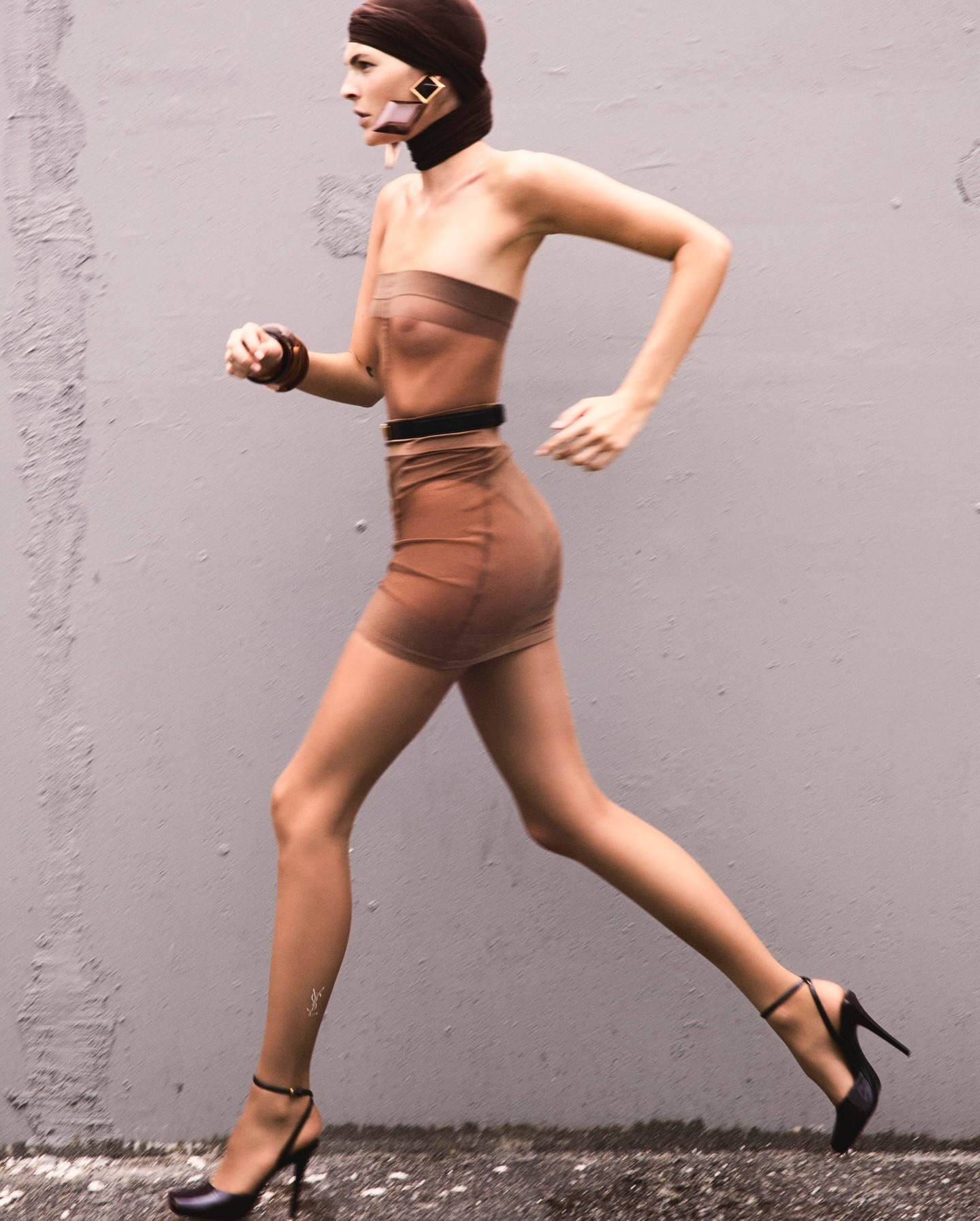 A woman in a brown strapless dress and high heels is in motion against a gray wall, wearing a head wrap and jewelry.