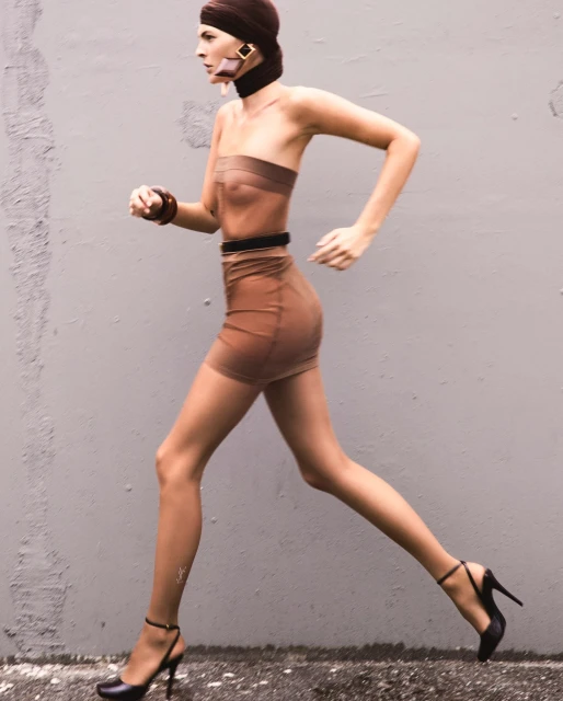 A woman in a brown strapless dress and high heels is in motion against a gray wall, wearing a head wrap and jewelry.