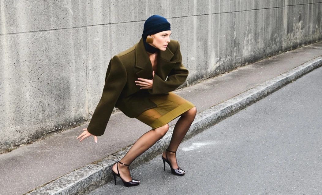 A woman in a fashion-forward olive jacket and skirt poses dramatically on a city street with a concrete wall in the background.