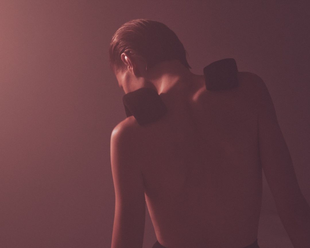 Back view of a bare-chested person with two dark rectangular objects on their shoulders against a softly blurred backdrop.