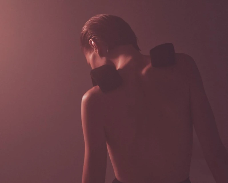 Back view of a bare-chested person with two dark rectangular objects on their shoulders against a softly blurred backdrop.