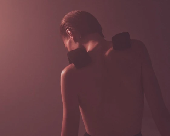 Back view of a bare-chested person with two dark rectangular objects on their shoulders against a softly blurred backdrop.