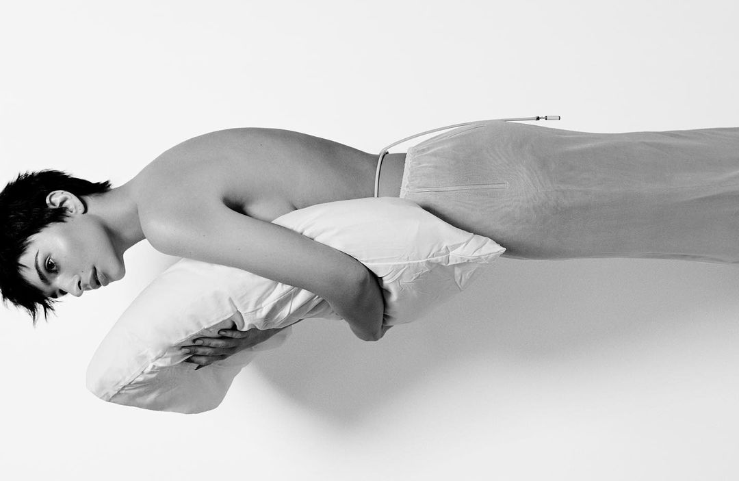 Monochromatic portrait of a light-skinned young adult lying on a white pillow, wearing a light-colored translucent garment, against a white background.