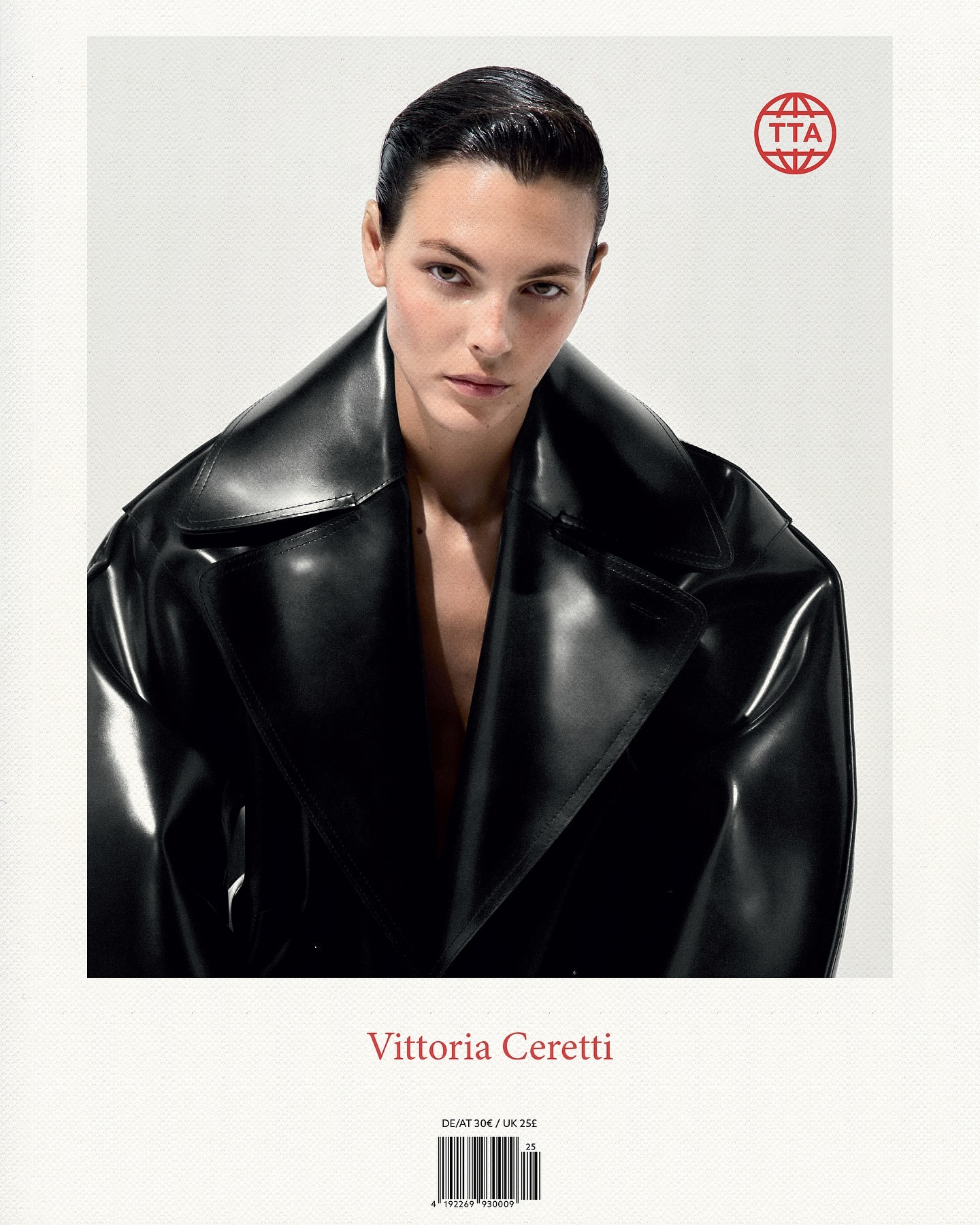 Close-up portrait of Vittoria Ceretti, with dark hair, wearing a shiny black trench coat against a bright, soft white background.