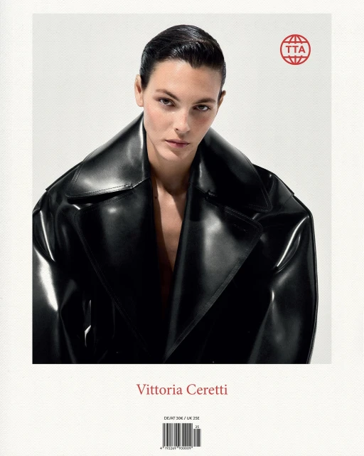 Close-up portrait of Vittoria Ceretti, with dark hair, wearing a shiny black trench coat against a bright, soft white background.