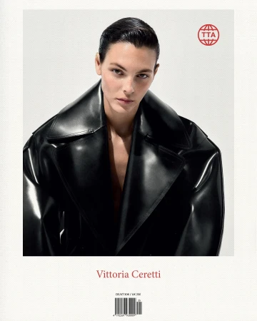 Close-up portrait of Vittoria Ceretti, with dark hair, wearing a shiny black trench coat against a bright, soft white background.