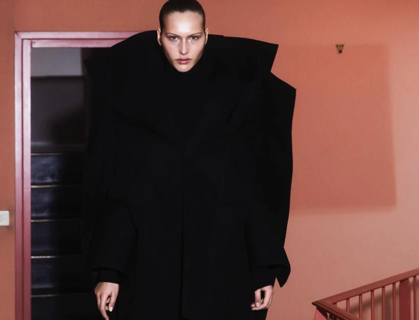 Woman in oversized black coat stands on stairs in front of a doorway. Dramatic fashion shot with neutral color scheme and artistic design.