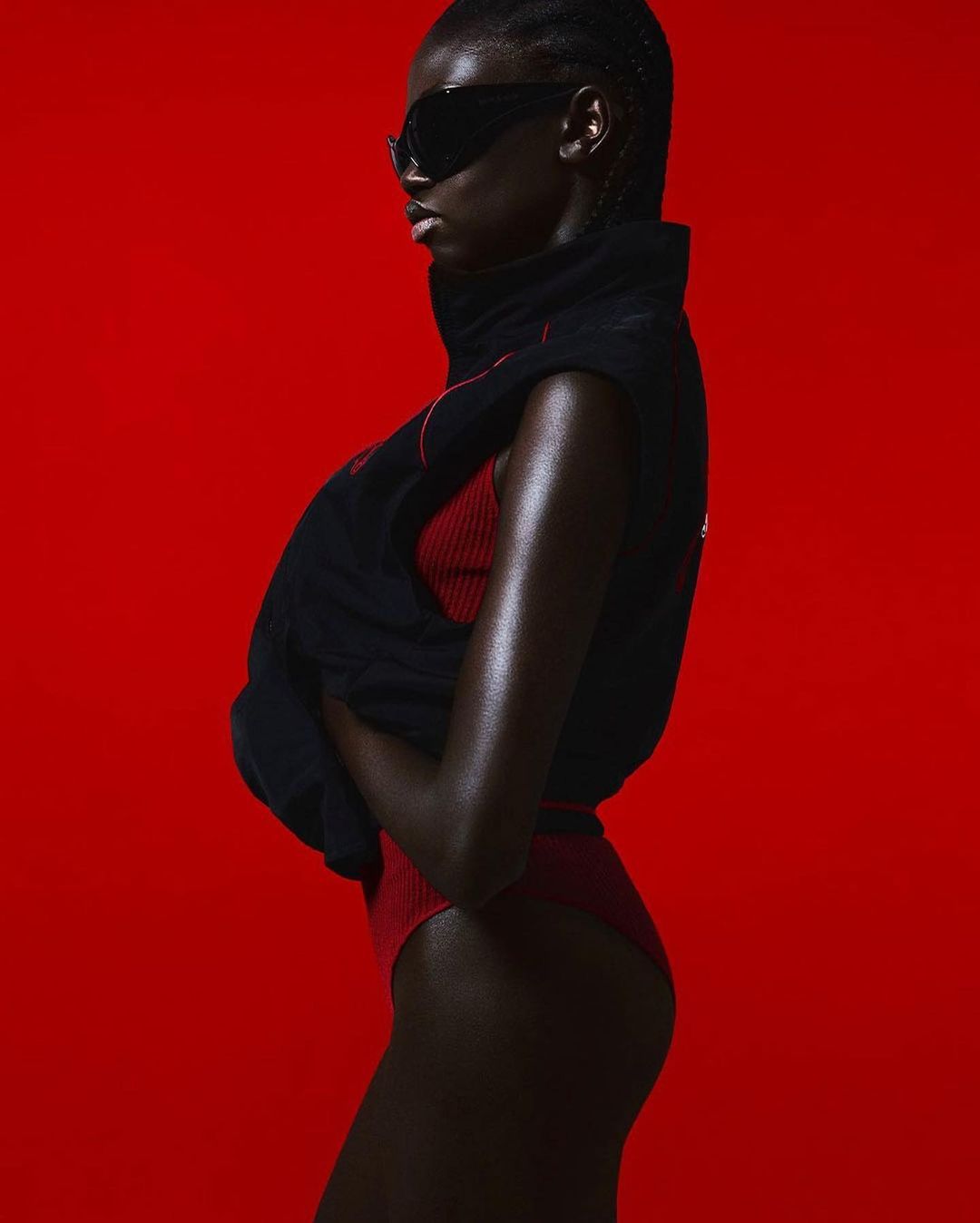 Fashionable woman in sunglasses posing against a vibrant red backdrop, wearing a red swimsuit and black vest in a studio setting.