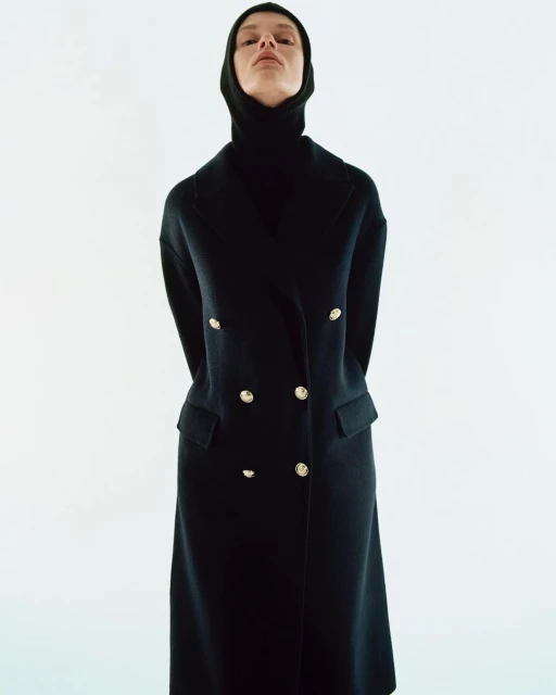 A woman wearing a long black double-breasted coat with gold buttons and a black hood stands against a white background in a fashion shot.