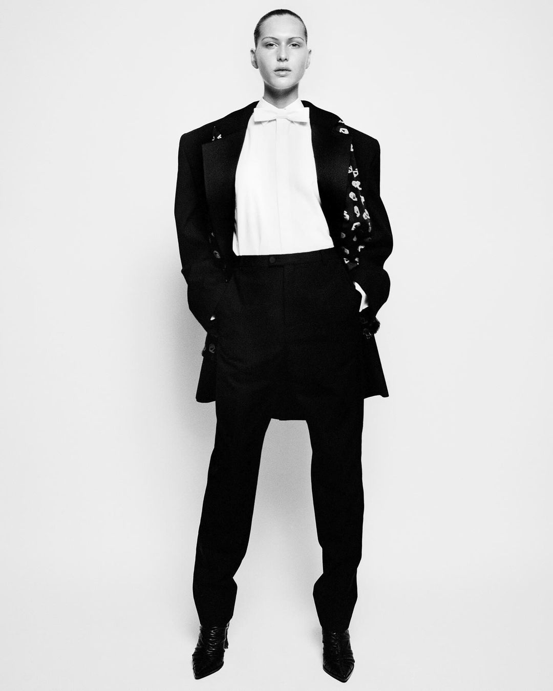 A fashion shot captures a solitary adult in an oversized black suit, crisp white shirt, and glossy black shoes against a bright backdrop.