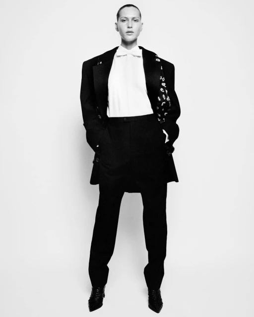A fashion shot captures a solitary adult in an oversized black suit, crisp white shirt, and glossy black shoes against a bright backdrop.