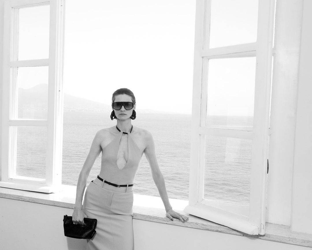 Model wearing dark sunglasses and skirt outfit holding a clutch while standing by large open windows overlooking the water.