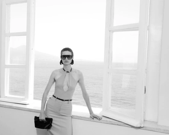 Model wearing dark sunglasses and skirt outfit holding a clutch while standing by large open windows overlooking the water.