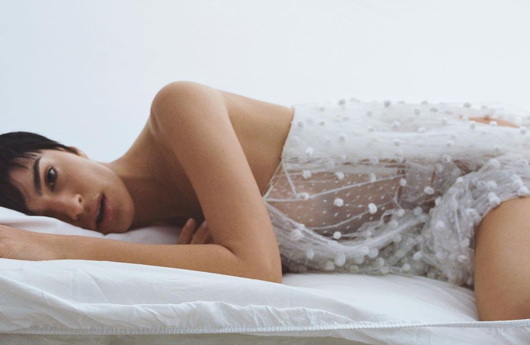 Portrait of a woman lying on a white sheet, partially covered by a sheer white dress with polka dots, looking at the camera.