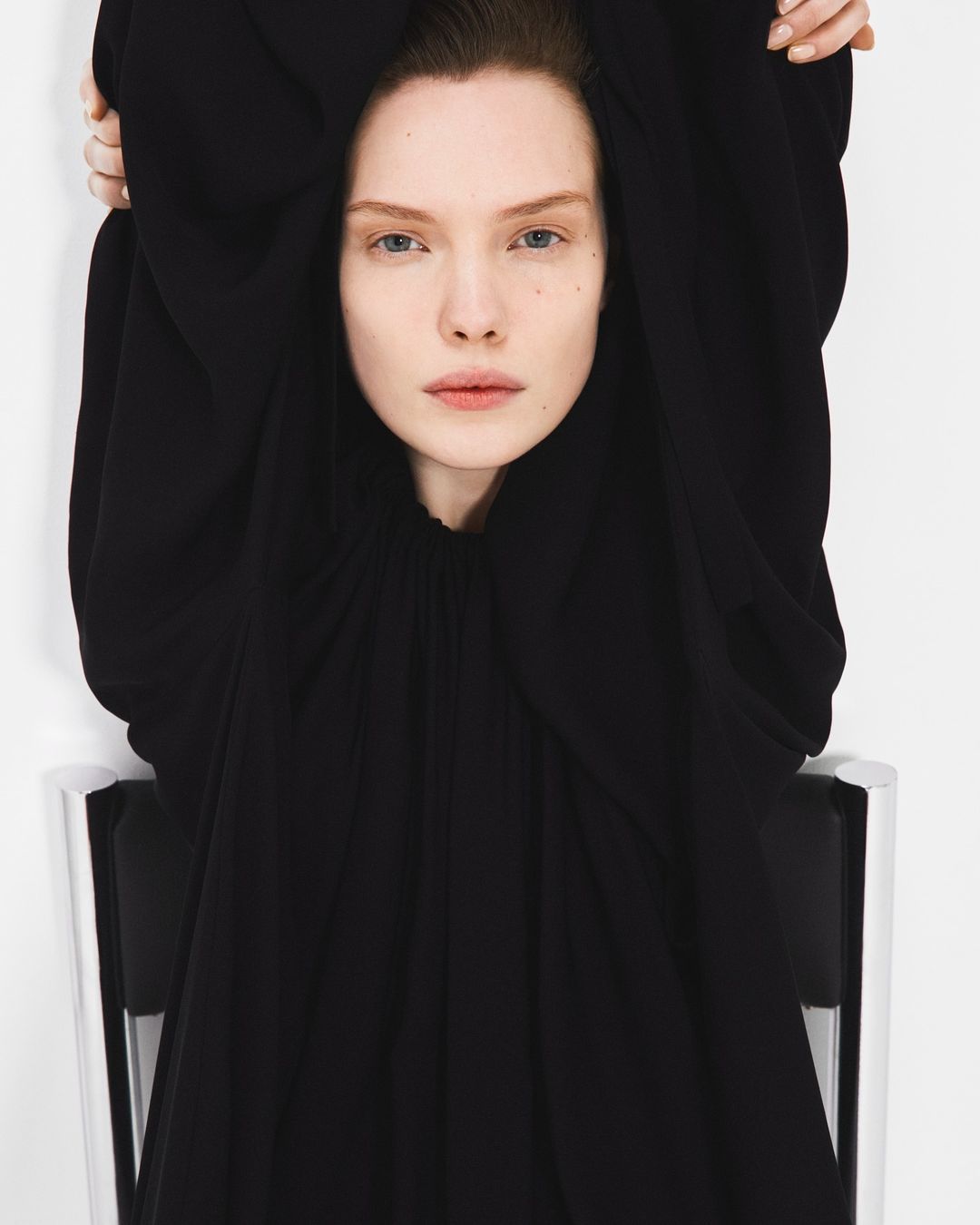 Portrait of a fair-skinned adult woman with dark hair, wearing a black top and sitting in a modern silver-and-black chair against a white background.