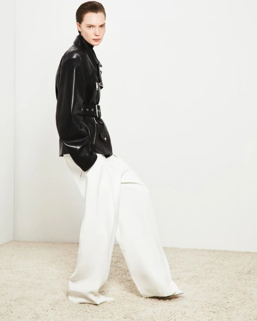 Fashion model poses in a black leather jacket and white wide-leg trousers against a white wall, showcasing a minimalist studio style.