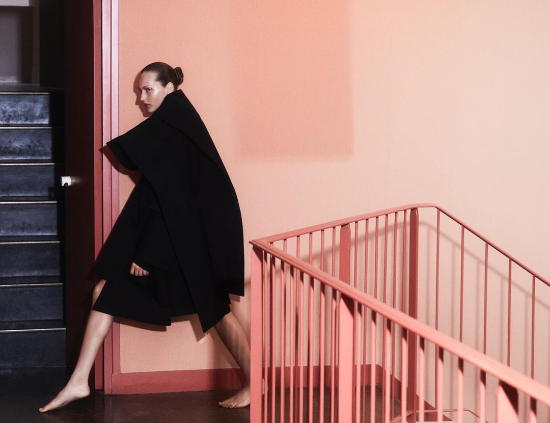 Woman in black cape poses on stairs with peach wall and pink railing, creating a striking fashion image with a minimalist aesthetic.