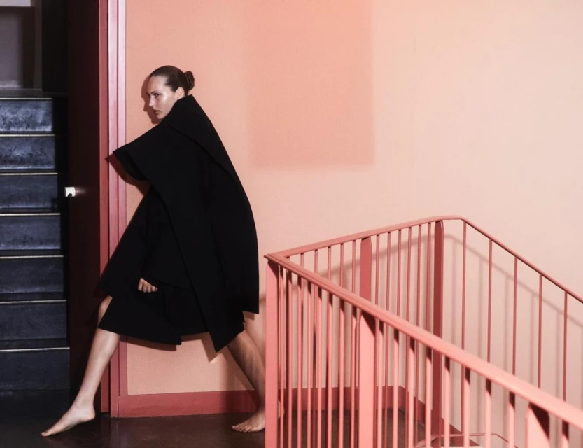 Woman in black cape poses on stairs with peach wall and pink railing, creating a striking fashion image with a minimalist aesthetic.