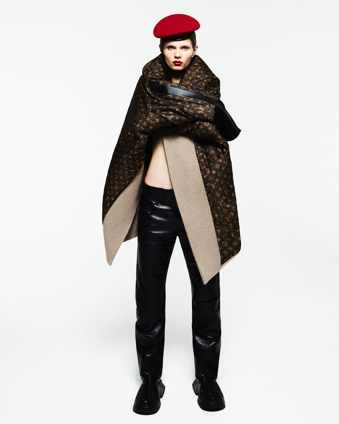 Fashion shot of a model wearing a red beret, a patterned brown cape, a black top, and shiny black pants against a white backdrop.