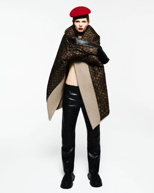 Fashion shot of a model wearing a red beret, a patterned brown cape, a black top, and shiny black pants against a white backdrop.