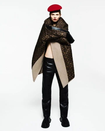 Fashion shot of a model wearing a red beret, a patterned brown cape, a black top, and shiny black pants against a white backdrop.