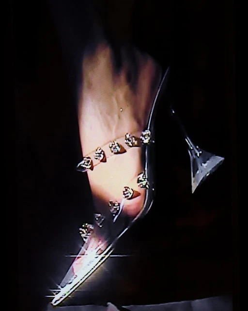 A woman's foot and ankle are adorned with a see-through, high-heeled shoe featuring jewel embellishments, set against a dark backdrop.