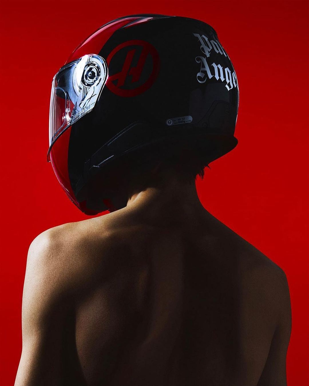 Back view of a person wearing a black and red Palm Angels motorcycle helmet against a vibrant red backdrop in a studio setting.