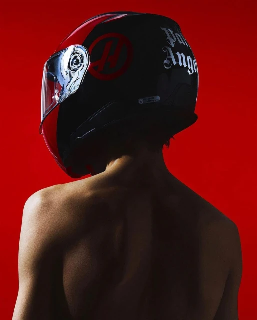 Back view of a person wearing a black and red Palm Angels motorcycle helmet against a vibrant red backdrop in a studio setting.