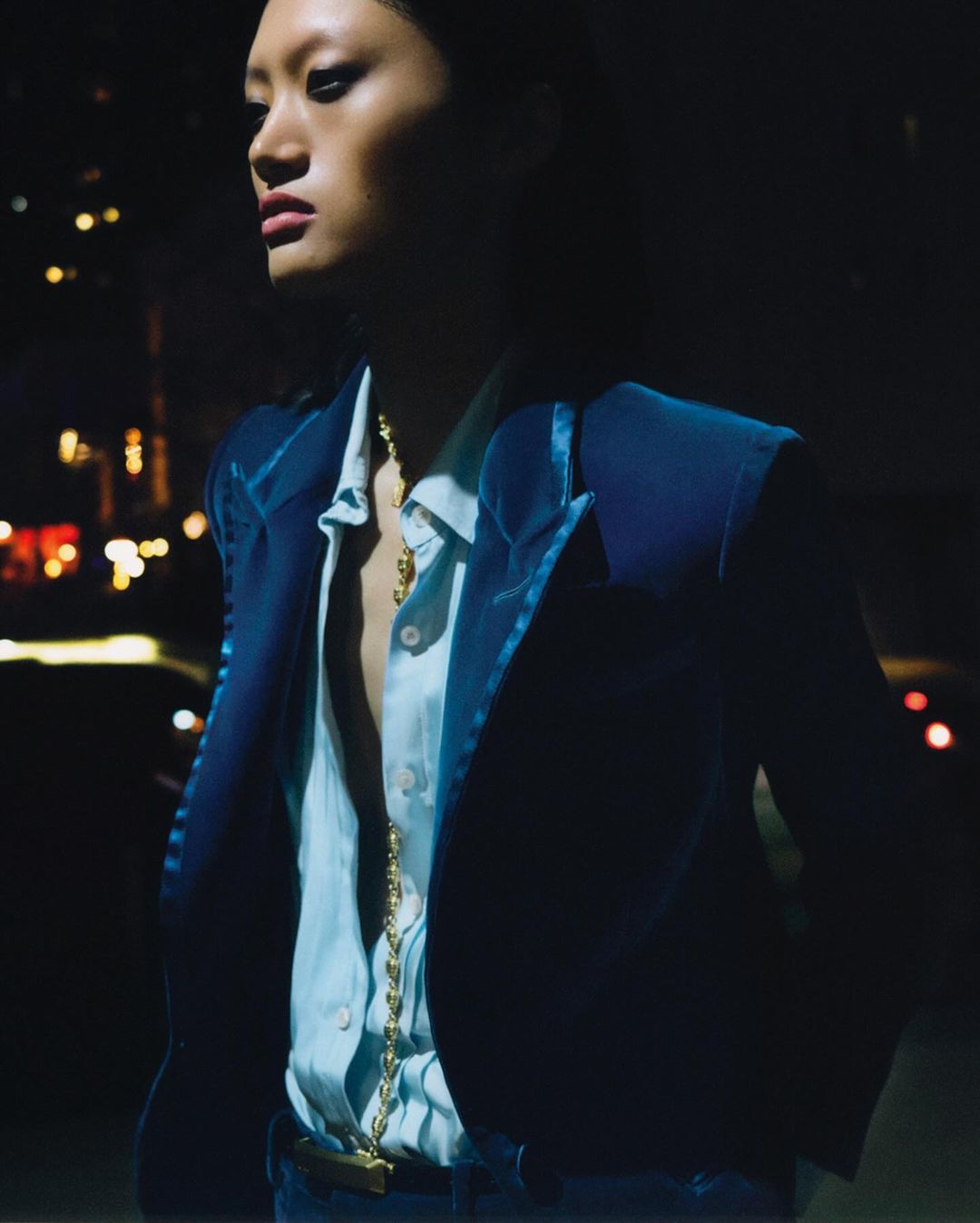 An Asian female model in a blue velvet jacket and unbuttoned light blue shirt poses at dusk, with bokeh lights in the background.
