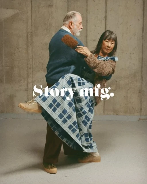 Elderly couple in casual wear in an embrace indoors in front of a concrete wall with neutral, soft lighting and visual brand text.