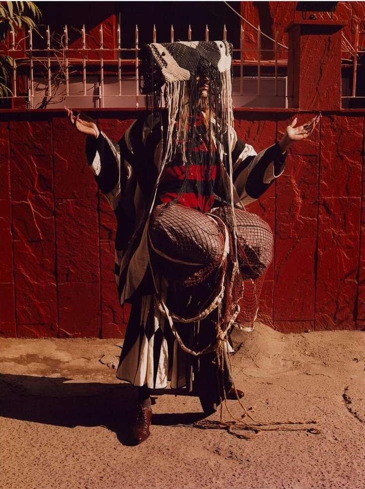 Person in patterned clothing with covered head standing in front of a red brick wall with a metal grate and a gray concrete ground.
