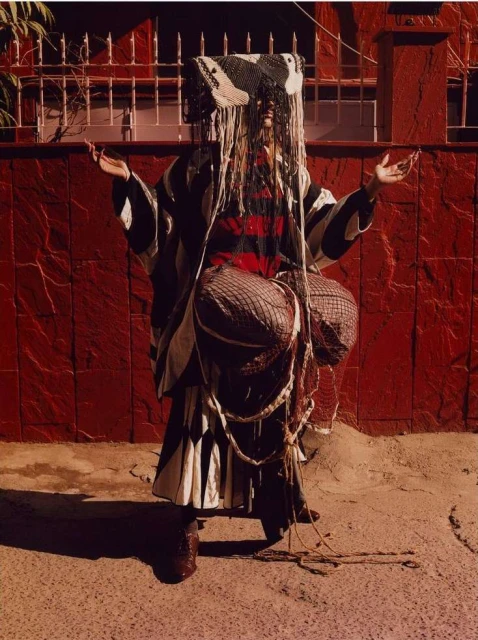 Person in patterned clothing with covered head standing in front of a red brick wall with a metal grate and a gray concrete ground.