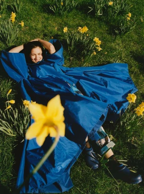 Woman in blue coat lies in daffodils smiling, surrounded by green grass. Outdoor shot, natural light, vibrant spring scene.