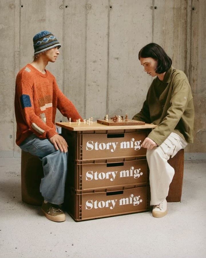 Two people play chess on a board placed on stacked storage boxes in an indoor setting with a concrete wall background and neutral colors.