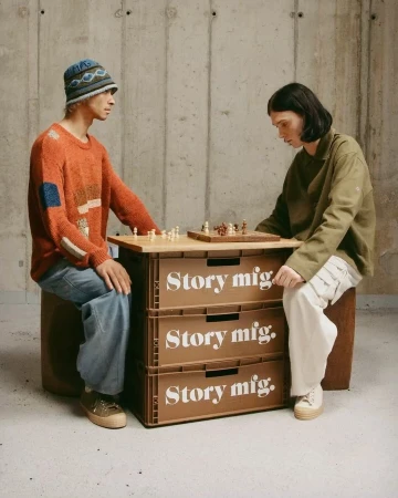 Two people play chess on a board placed on stacked storage boxes in an indoor setting with a concrete wall background and neutral colors.