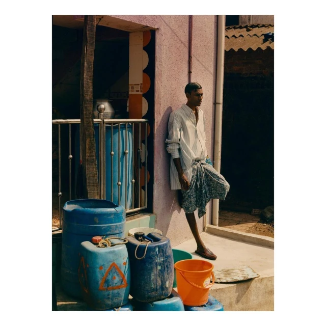 Man in casual white shirt and printed wrap skirt leans against a pink wall next to blue water containers outdoors on sunny day.
