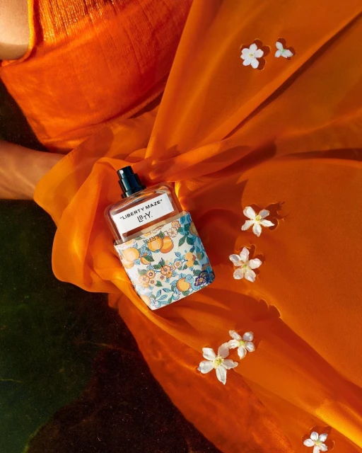 Perfume bottle with orange patterned cover, surrounded by white flowers on an orange silk background, flat lay composition.