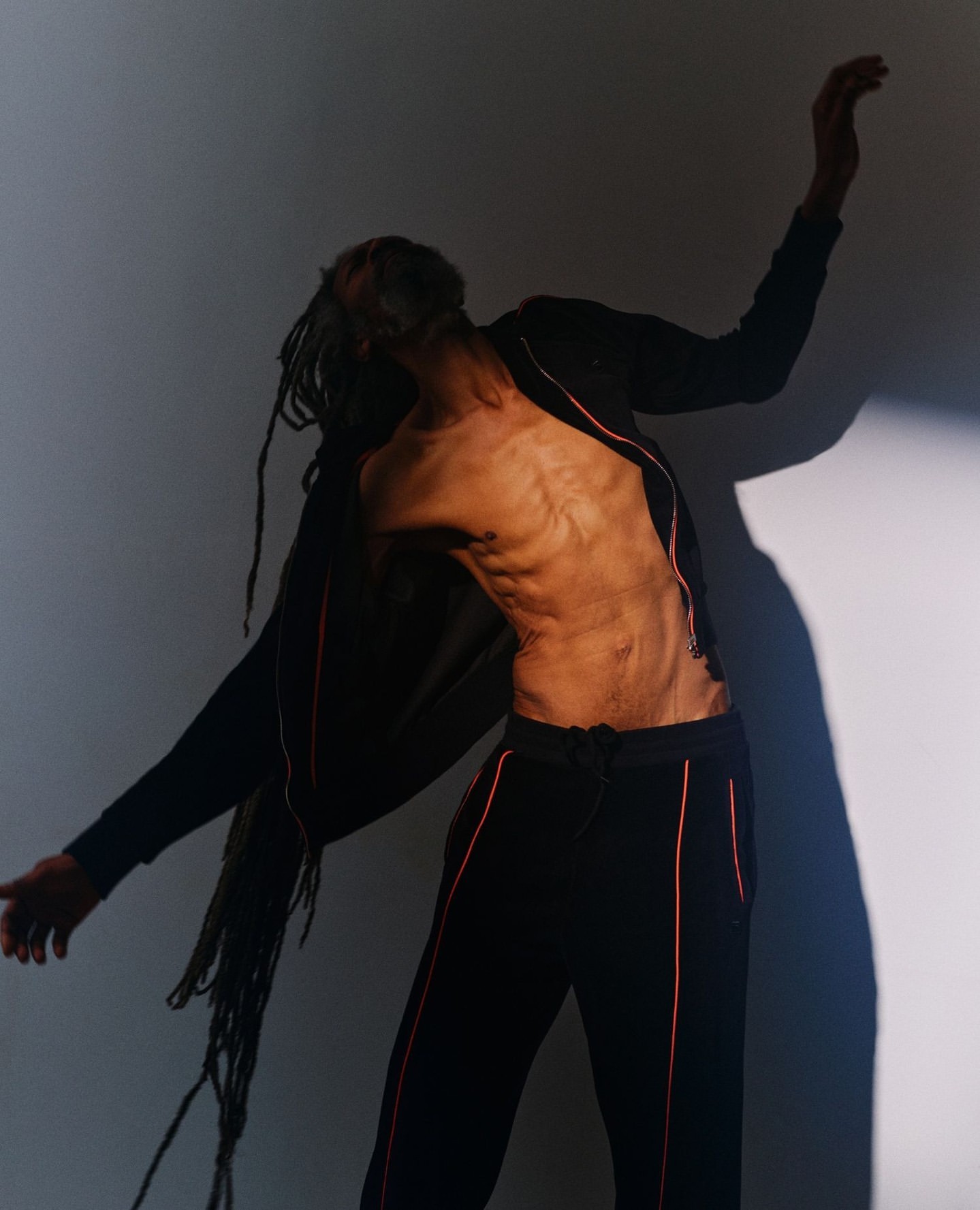 Artistic studio shot of a person with long dreadlocks wearing athletic clothes and posing against a neutral backdrop with dramatic lighting.