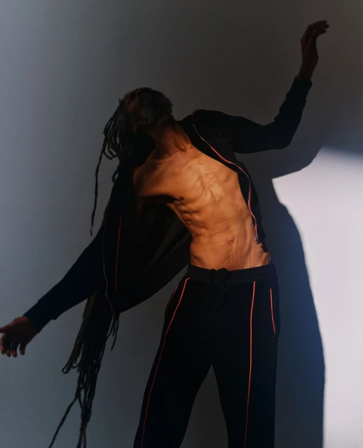Artistic studio shot of a person with long dreadlocks wearing athletic clothes and posing against a neutral backdrop with dramatic lighting.