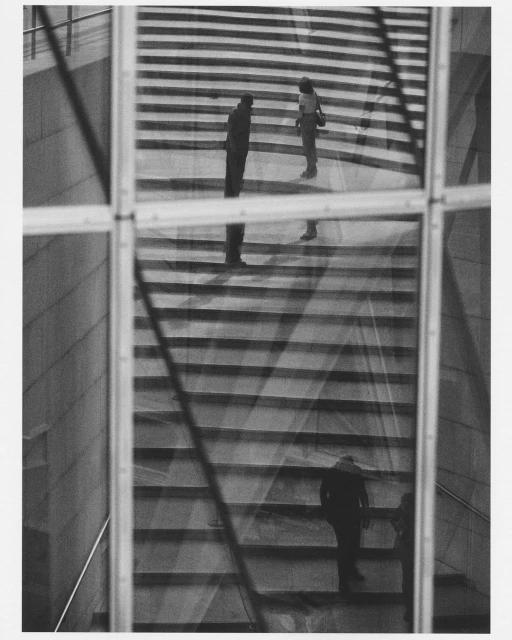 Black and white photograph featuring people on stairs, viewed through glass panels, creating a layered, geometric composition.