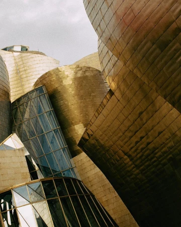 Close-up architectural image of the Guggenheim Museum Bilbao with geometric forms, titanium cladding, and glass features under a cloudy sky.