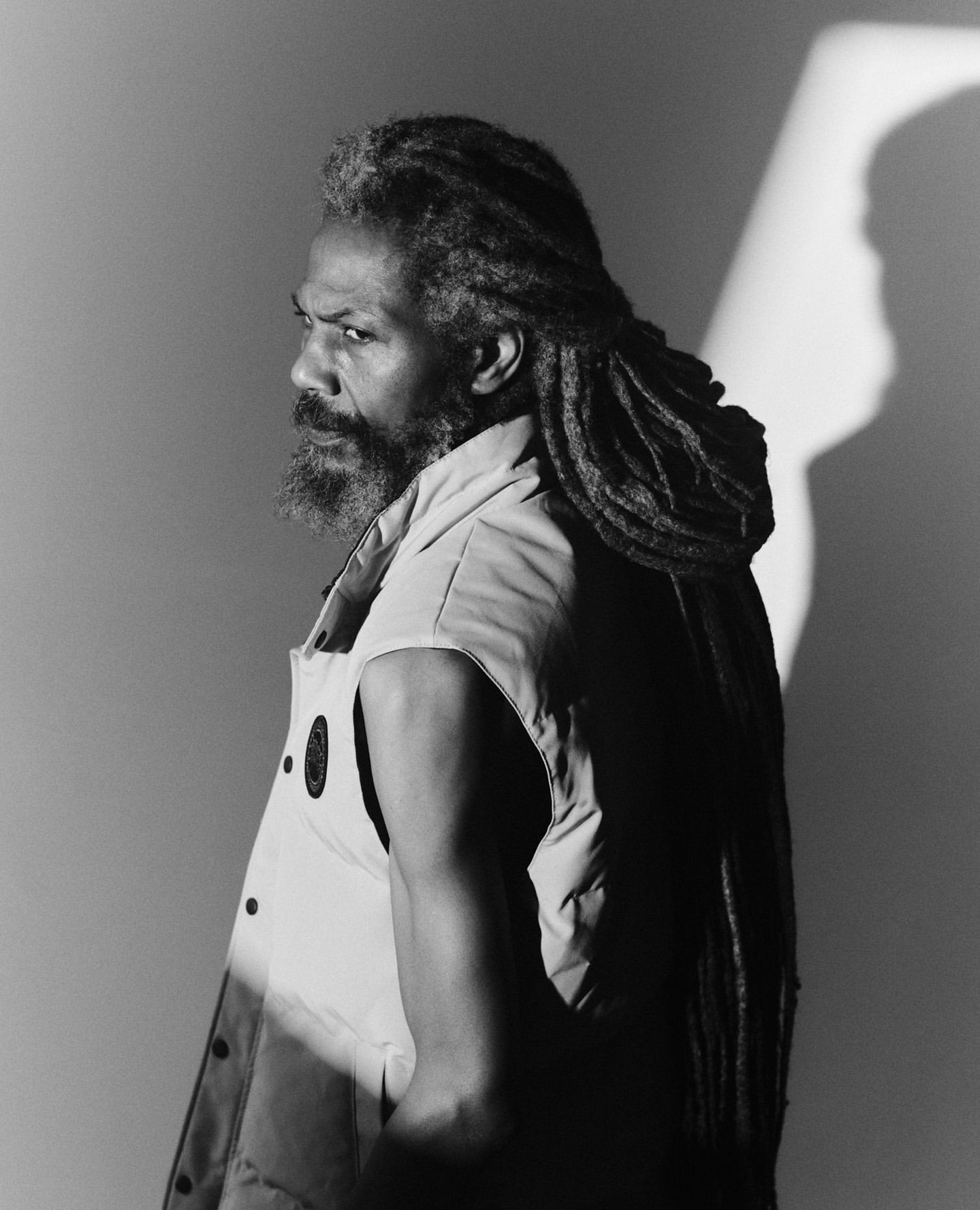 Black and white portrait of a Black man with dreadlocks and a beard, looking over his shoulder in a sleeveless jacket with a white background.